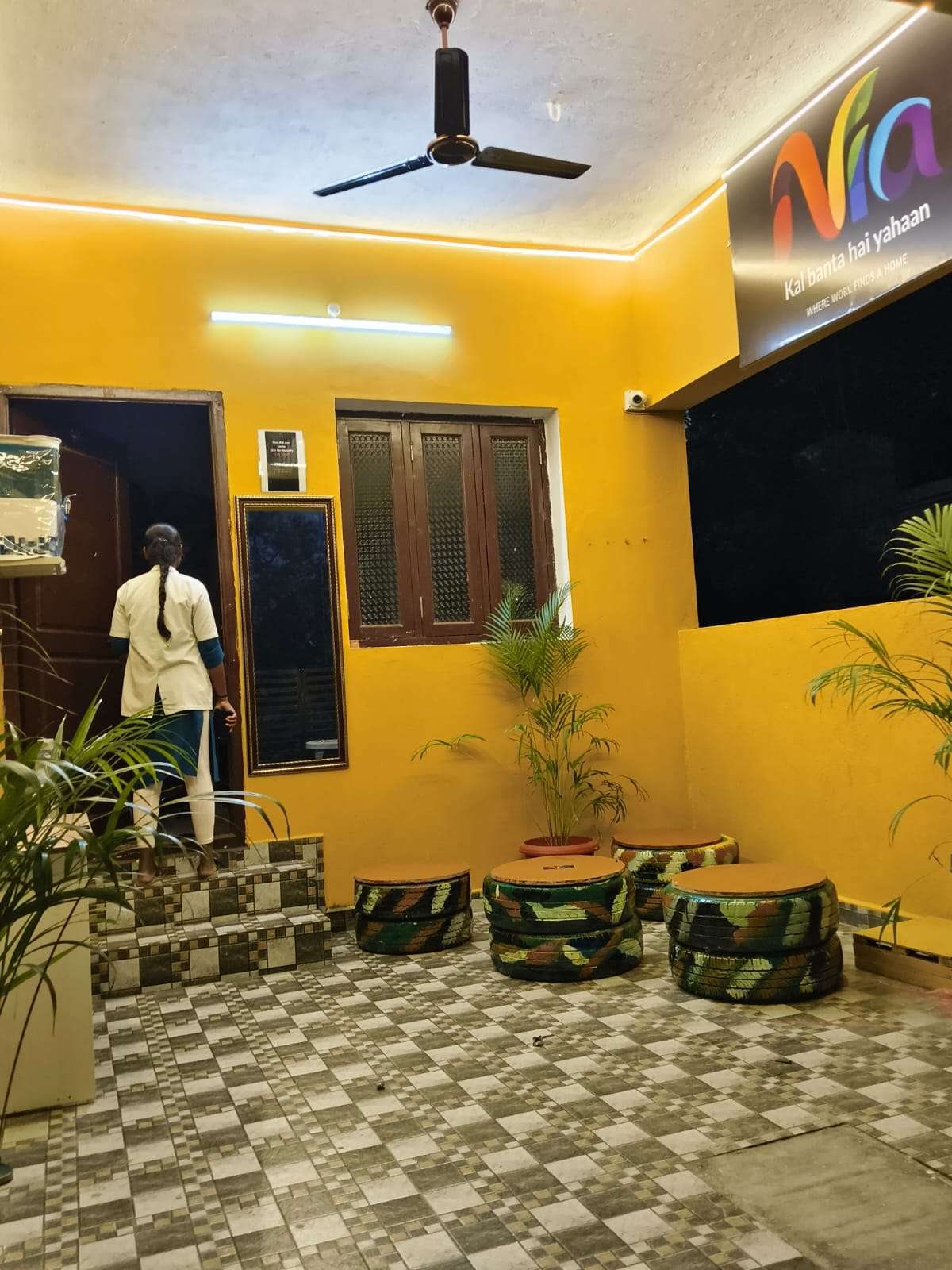A Nia Studio lounge at night: yellow walls, plants and seats made from painted tyres under a Nia sign